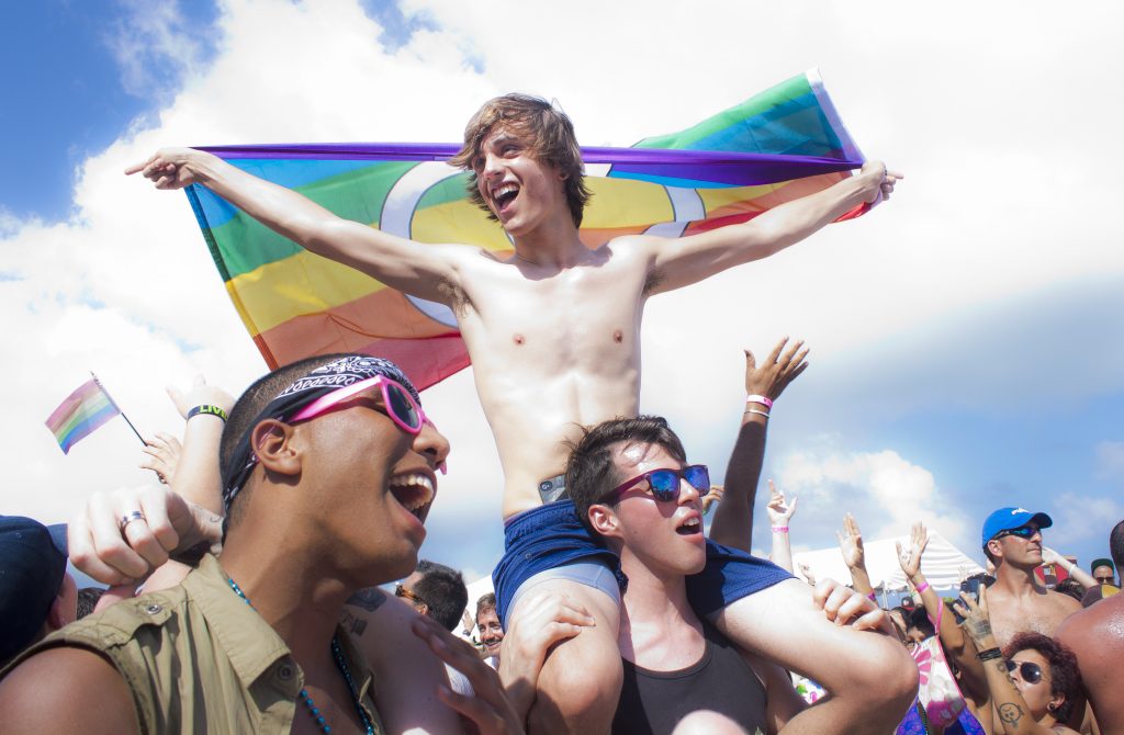 A young man holding up the pride flag while on the shoulders of a friend.